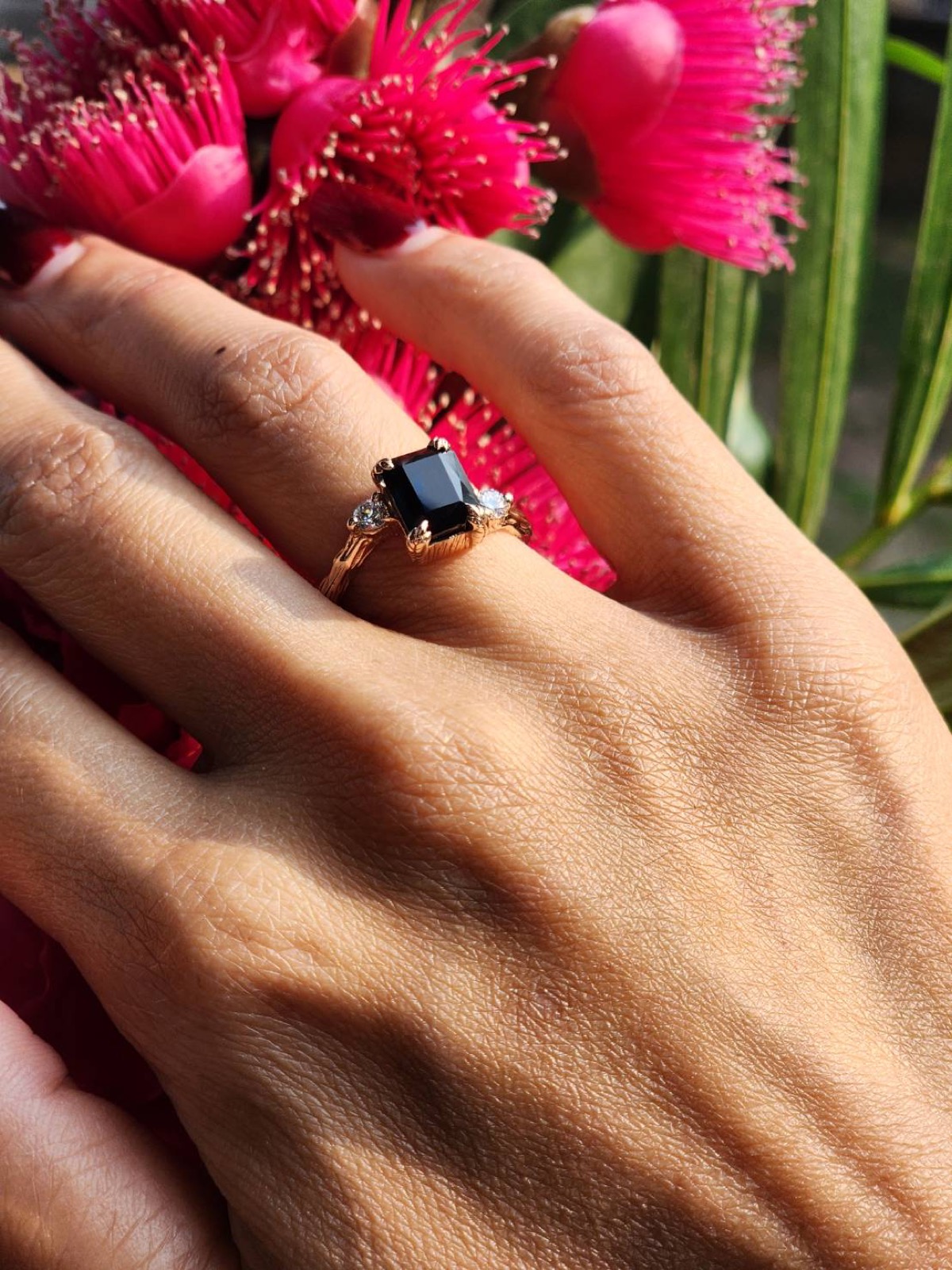 A finished dark gemstone ring worn outdoors beside vivid flowers.