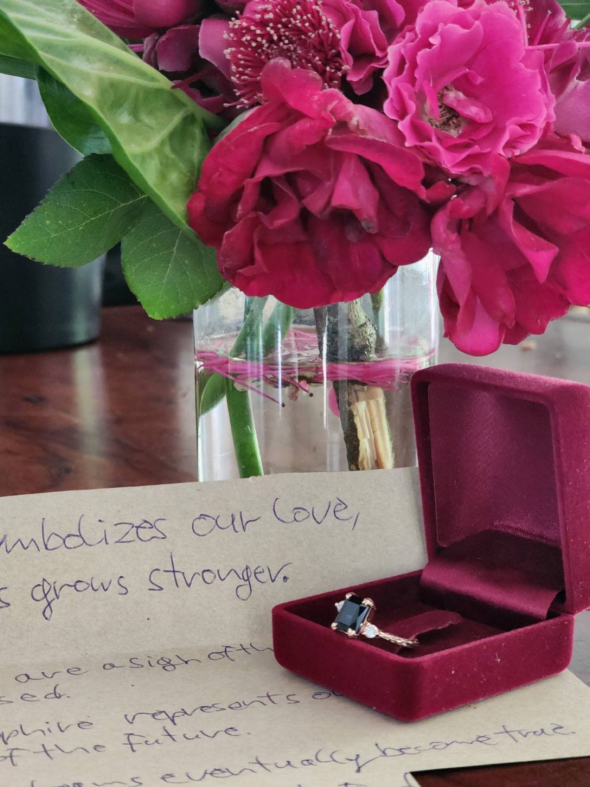 A finished gemstone ring presented in a velvet box beside flowers and a handwritten note.