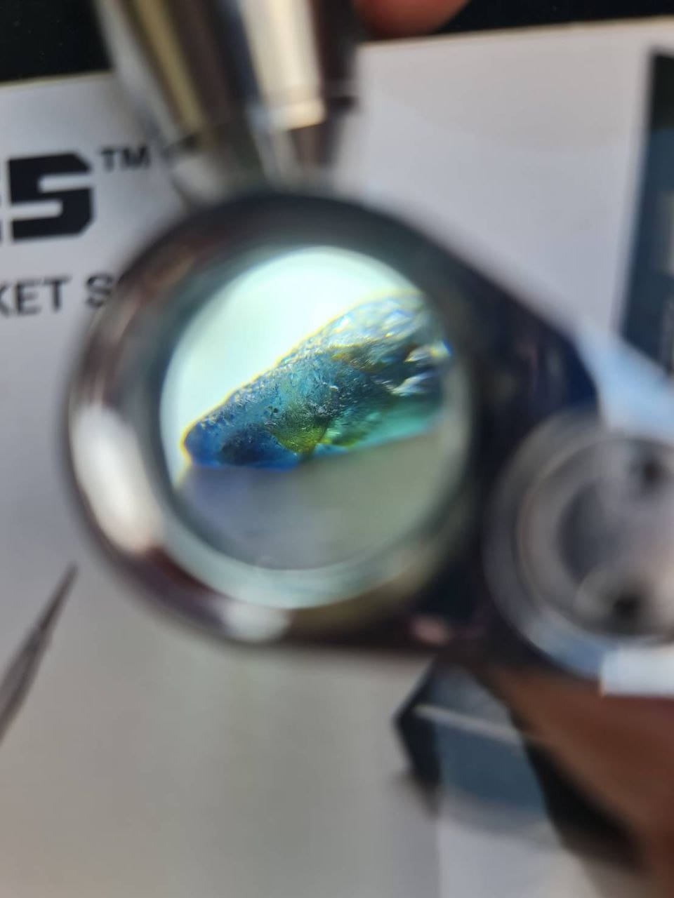 A rough blue-green gemstone magnified under a jeweler's loupe.