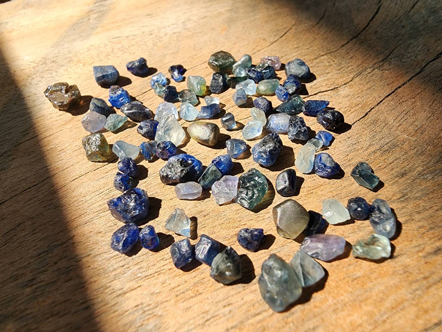 Mixed rough blue, green, and pale gemstones spread in sunlight on a wooden surface.