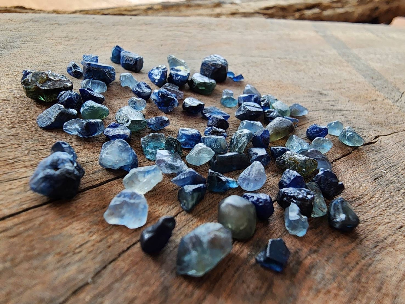 Many rough blue and green sapphires spread across a wooden surface.